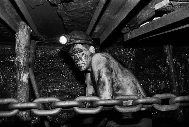 Black and white photo of a shirtless miner in a coal mine, black with coal wearing lighted helmut, chain in foreground.